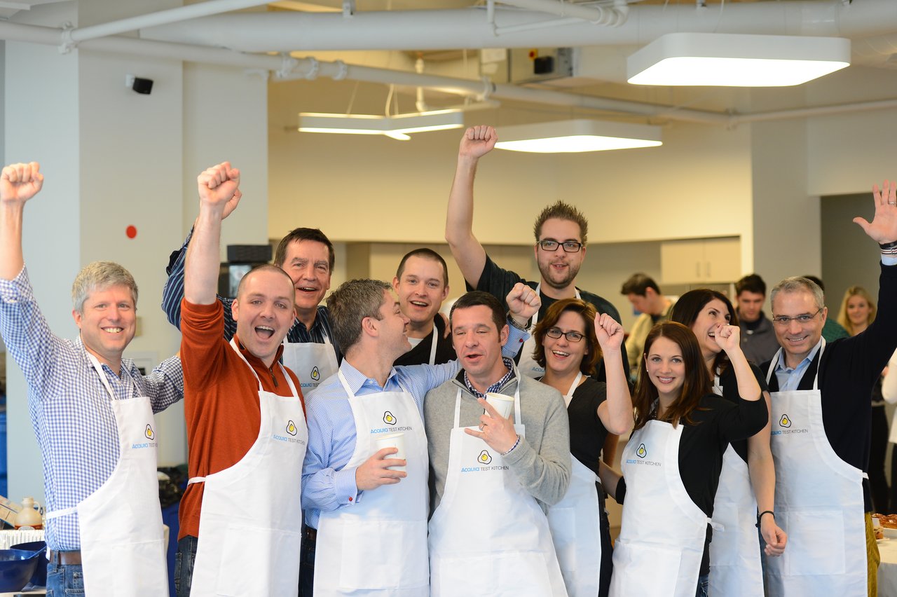 A group of people wearing aprons cheer and raise their fists in a kitchen setting.