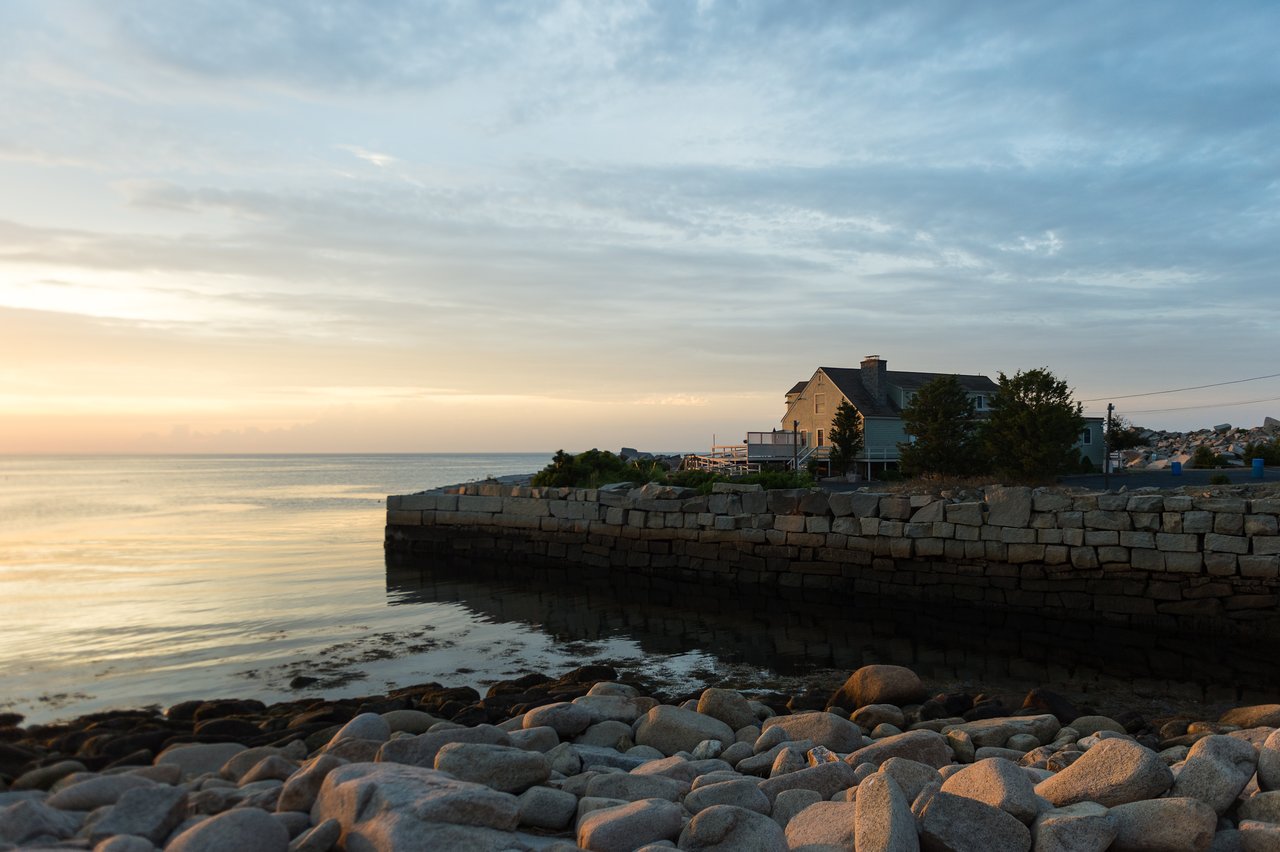A stone-lined harbor with calm water, a house in the background, and a rocky shoreline at sunset.