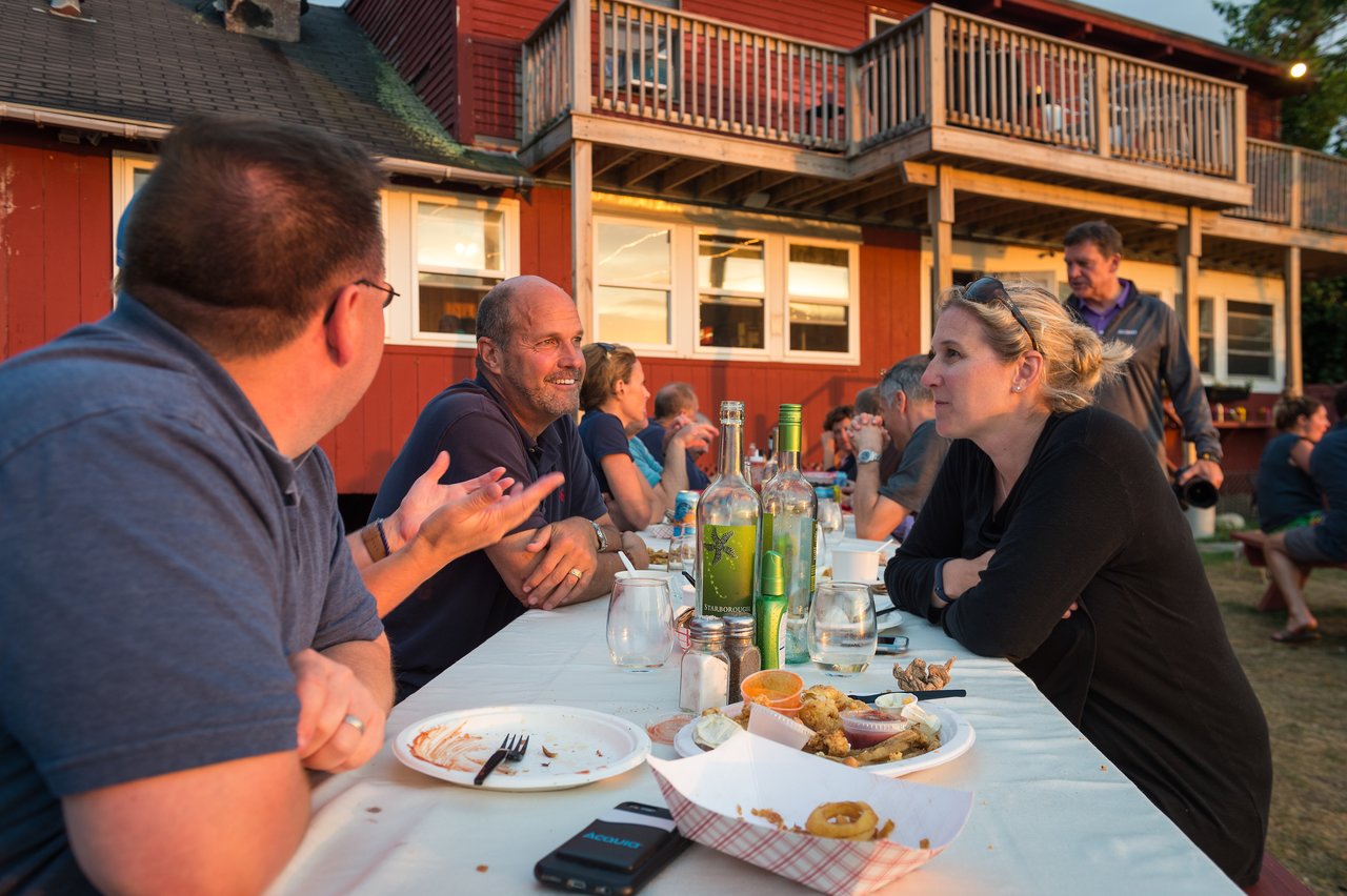 People sit at an outdoor table, eating and talking, with plates of food and drinks in front of them.