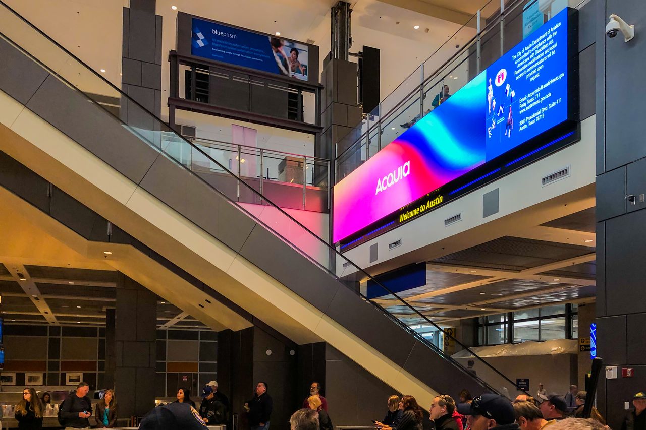 A large digital billboard in an airport displays "Acquia" with colorful graphics and a "Welcome to Austin" message.