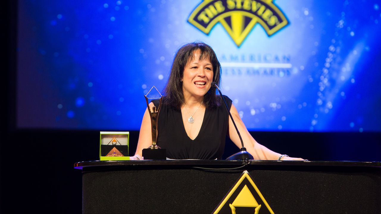 A woman smiles while holding a trophy and speaking at the Stevie Awards ceremony.