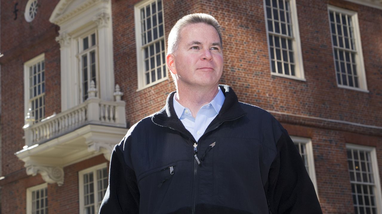 A man in a black jacket stands outside a brick building, looking into the distance.