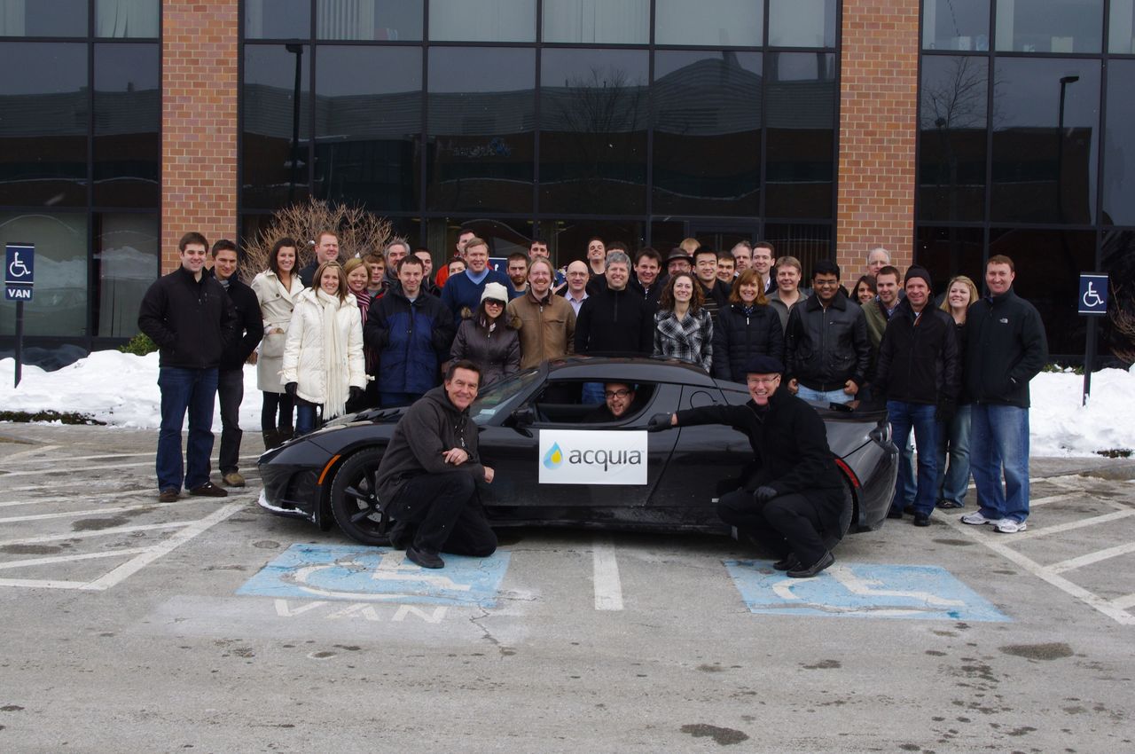 A group of people poses around a black Tesla Roadster with an "Acquia" sign on the side.