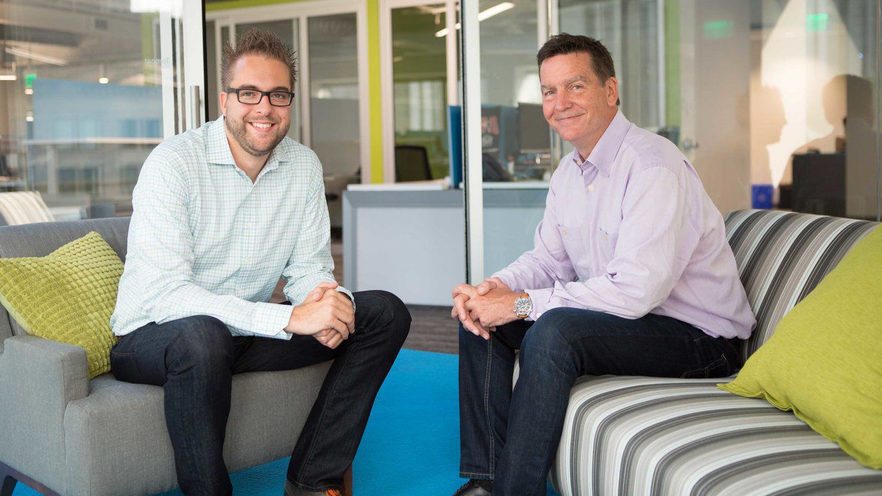 Two men sitting in an office lounge, smiling at the camera.