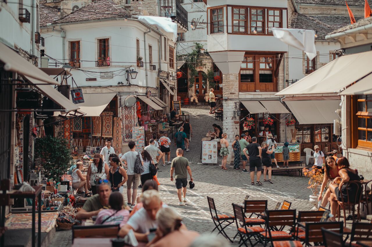 Tourists stroll and sit at cafes on a cobblestone street lined with shops in an old town.