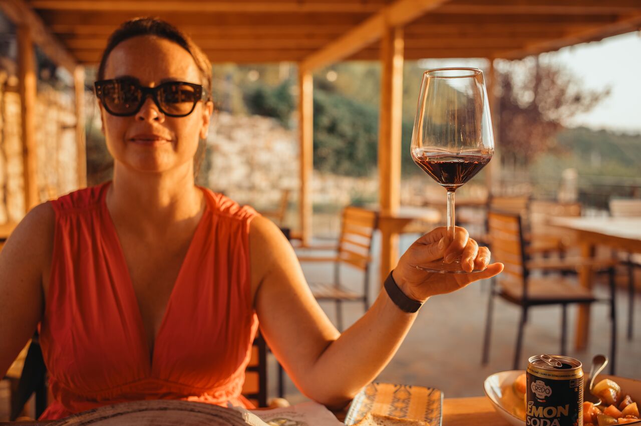 Woman in a red dress and sunglasses holding up a glass of red wine at an outdoor restaurant during golden hour.