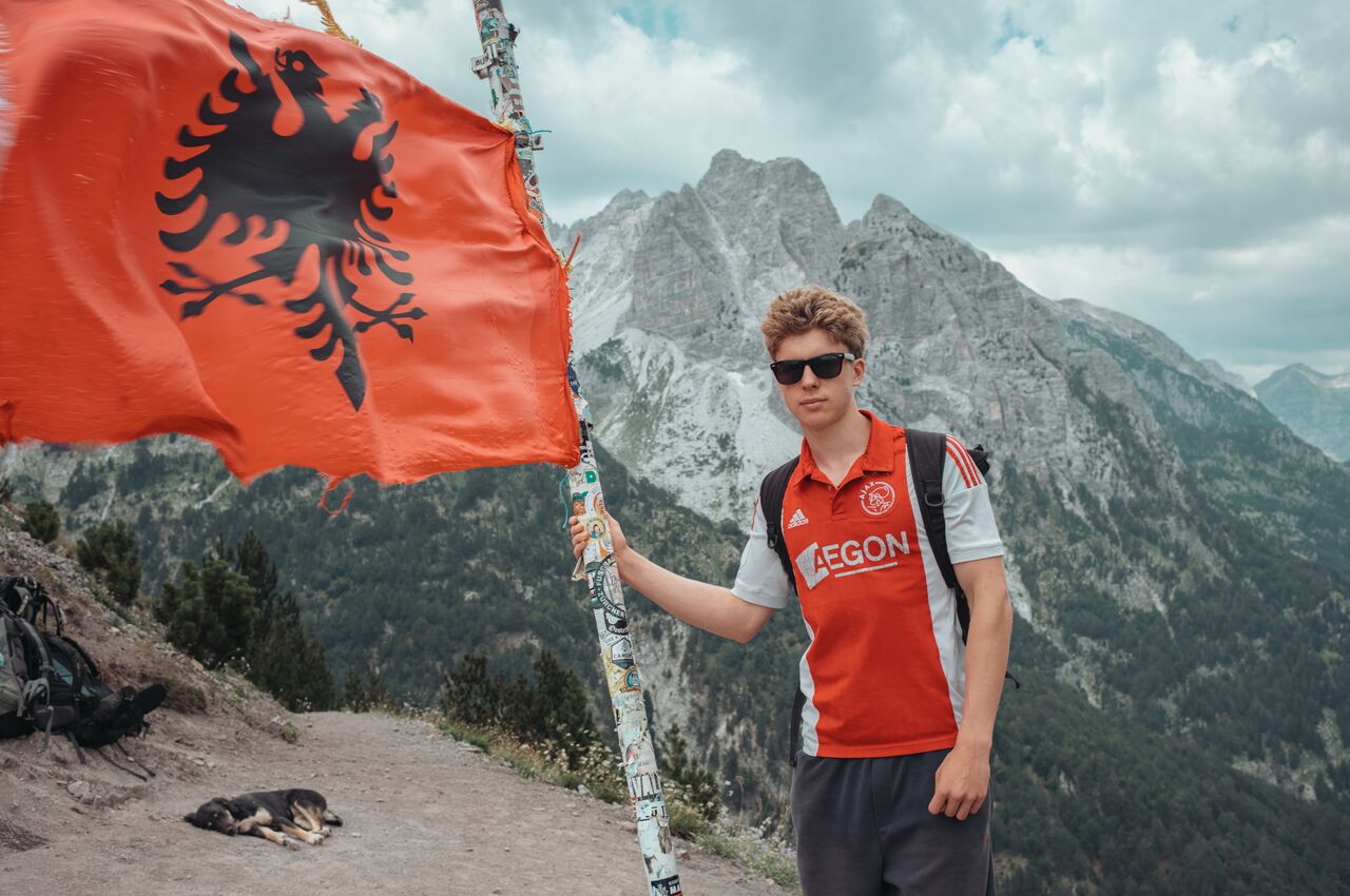A hiker holds a pole flying the Albanian flag at a mountain pass, with jagged peaks behind him.
