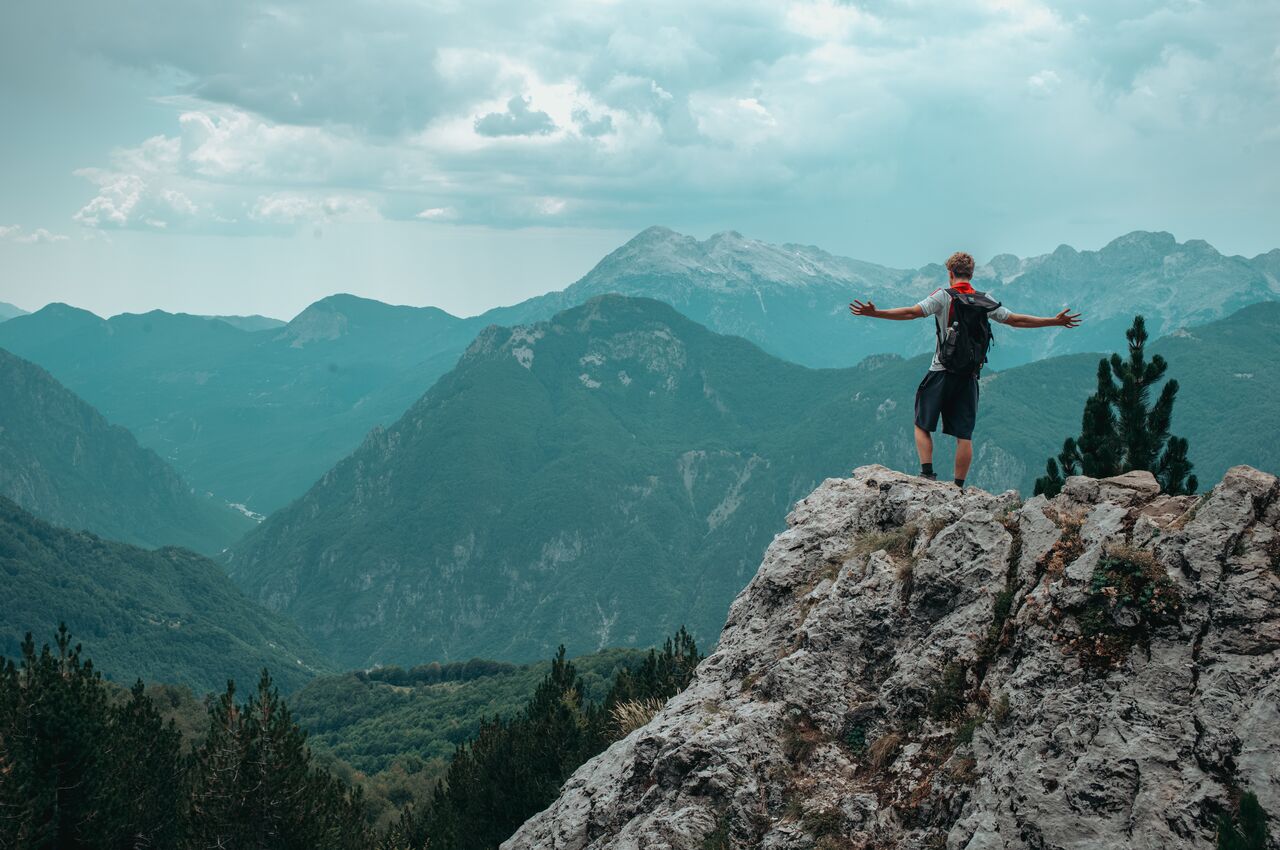 A hiker with a backpack stands on a rocky peak with arms outstretched, facing a vast range of green and gray mountains.