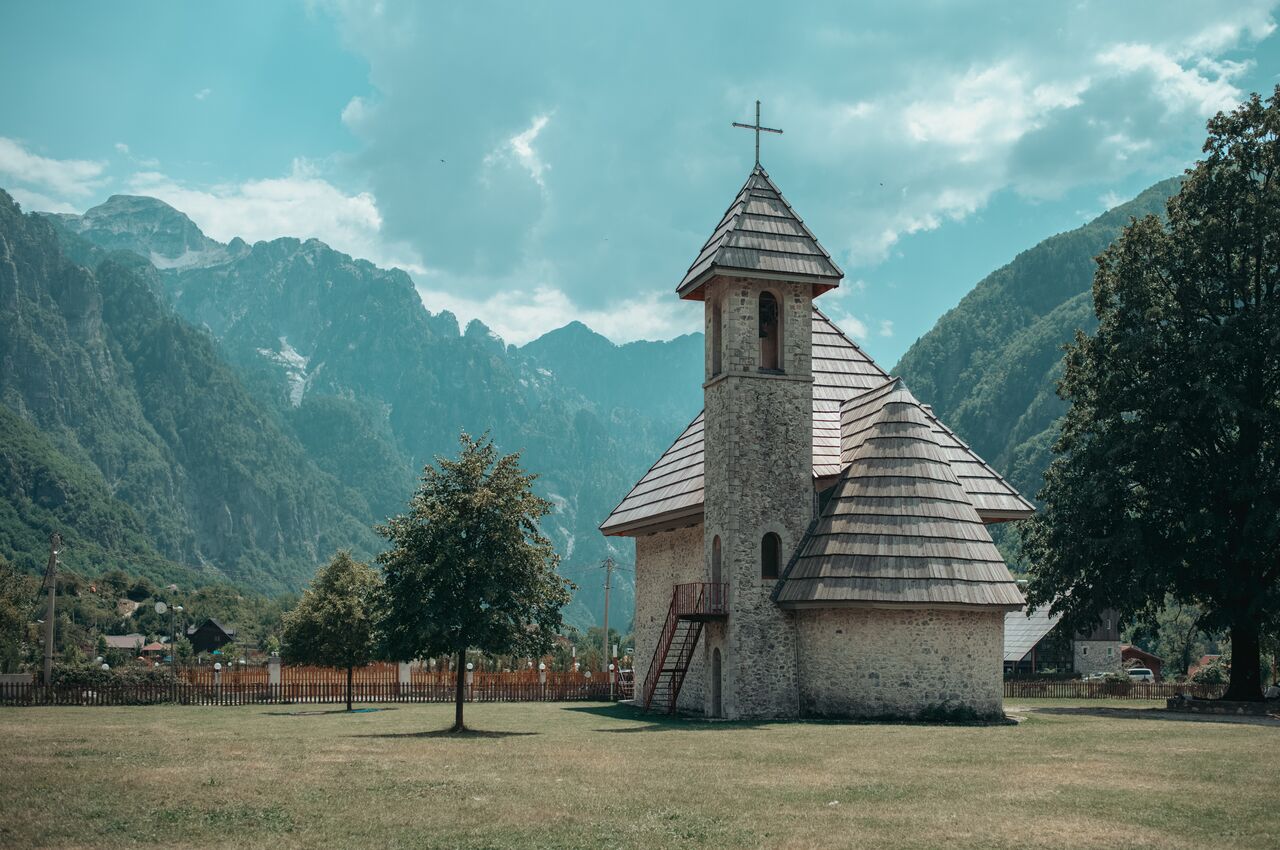 A small stone church with a wooden shingle roof and cross-topped steeple set against rugged mountains.
