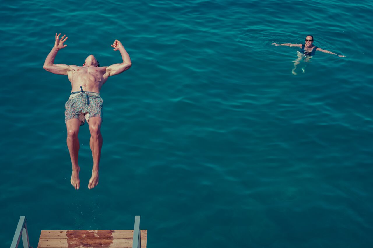 A man leaps off a wooden dock into the ocean as a woman in sunglasses swims nearby watching.