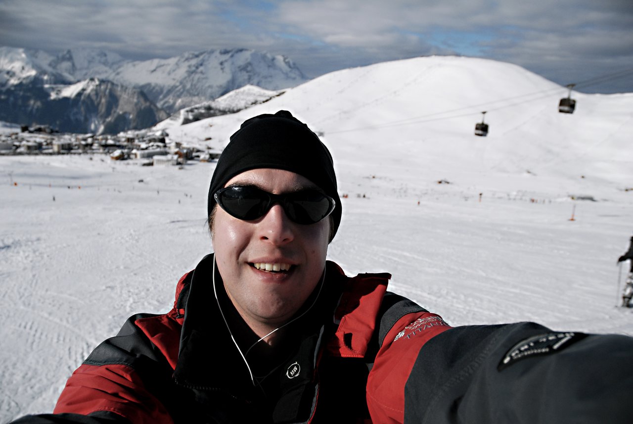 A person wearing sunglasses and a winter jacket takes a selfie on a snowy mountain with ski lifts in the background.