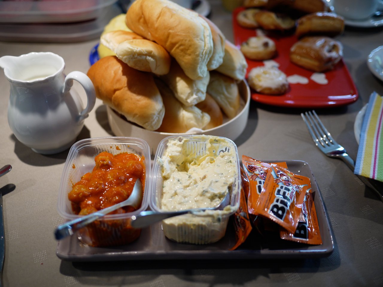 A table set for lunch with bread rolls, spreads, jam, and packets of sauce on a tray.