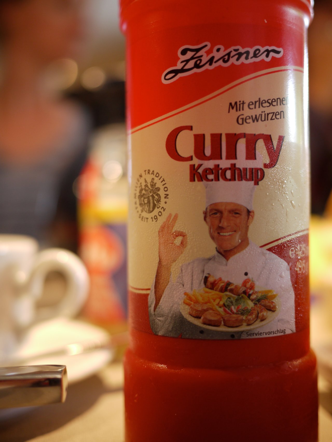 A bottle of curry ketchup is in focus on a table with plates, cups, and food in the background.