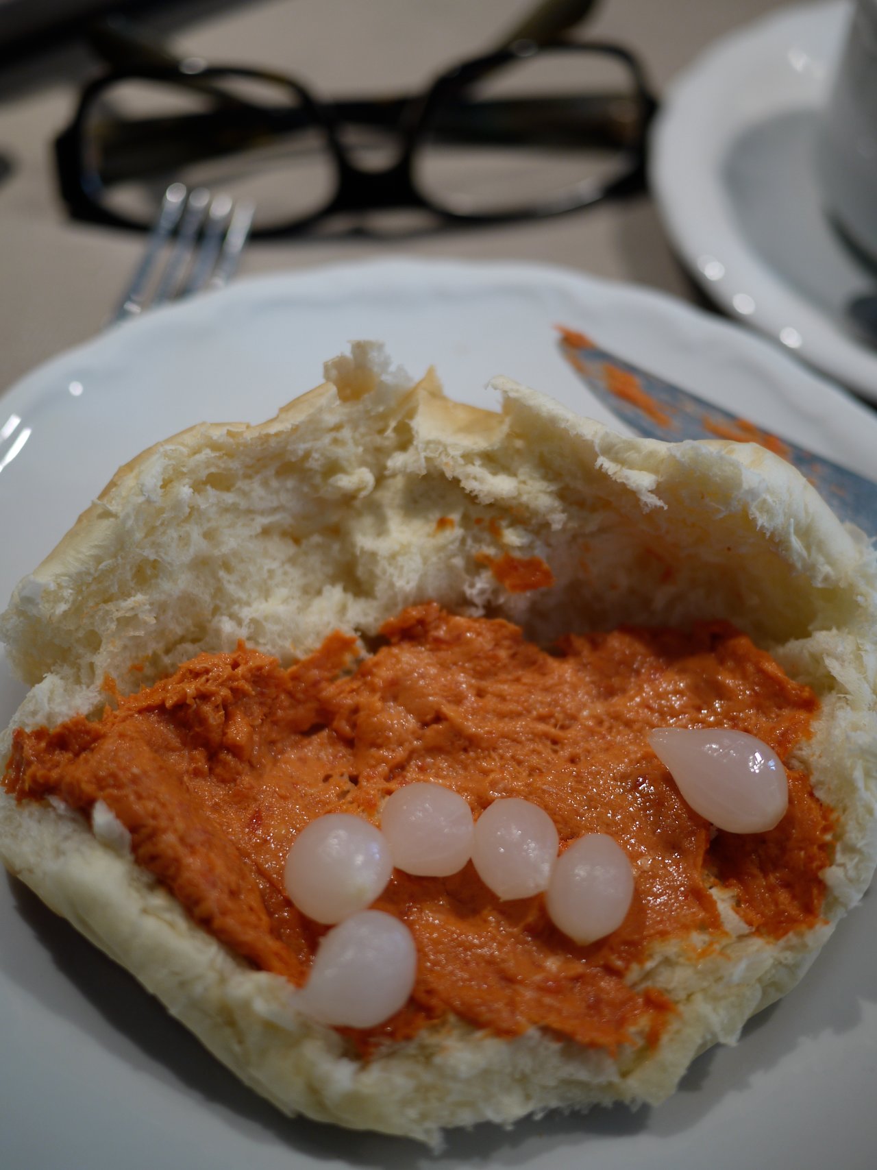 A bread roll with orange spread and small white pearl onions on a plate, with a knife and glasses nearby.