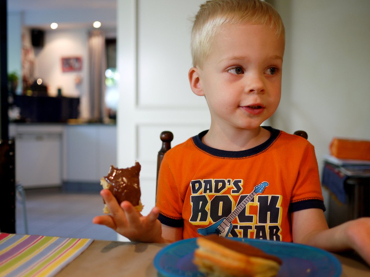 A young child in an orange shirt holds a piece of bread with chocolate spread while sitting at the table.