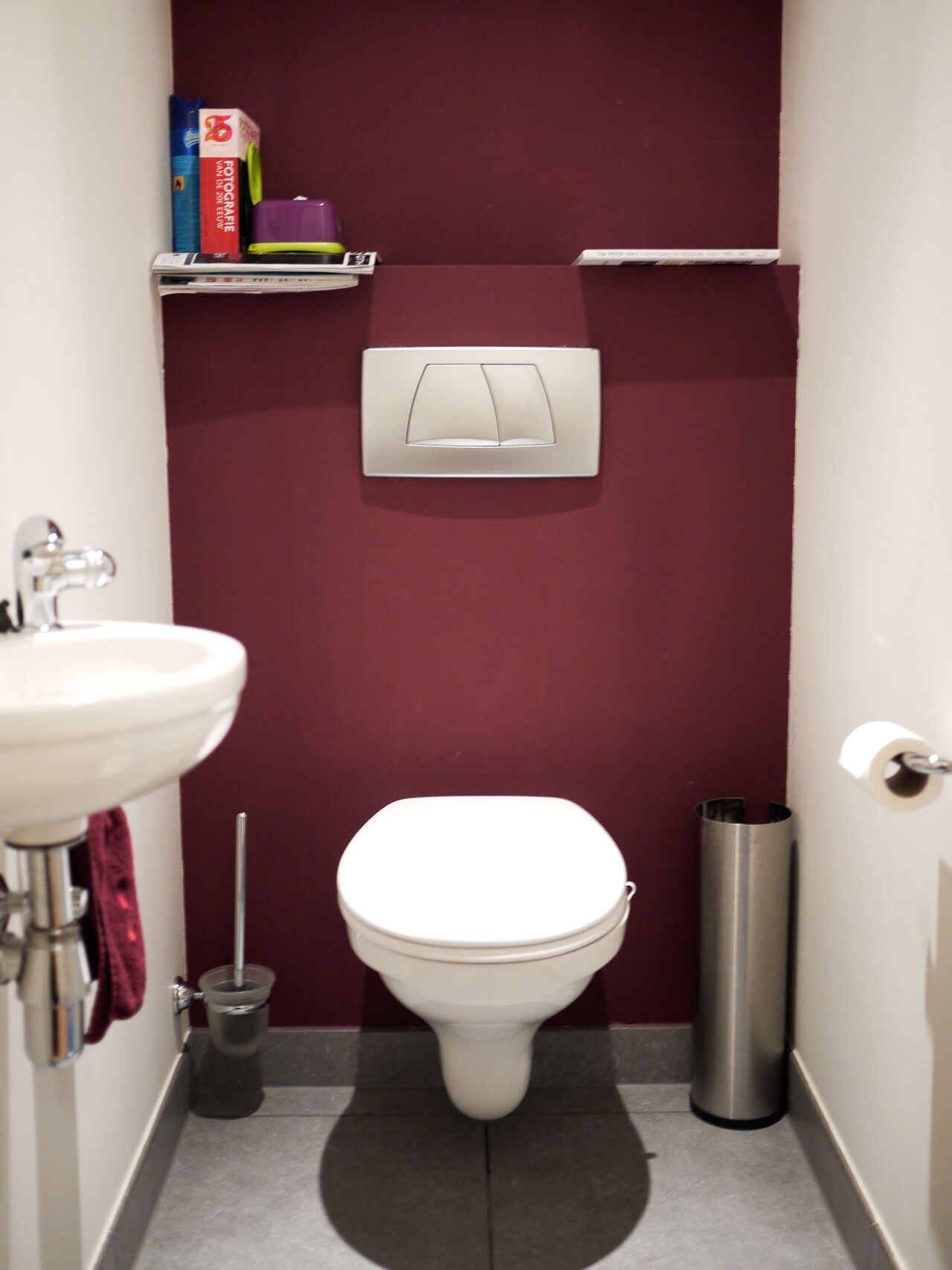 Small bathroom with a wall-mounted toilet, sink, and shelves holding toiletries against a dark red wall.