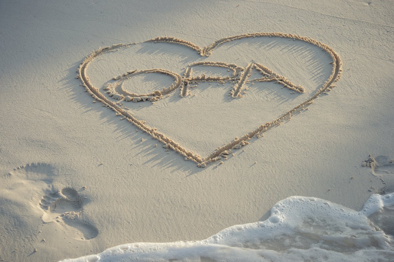 A heart with the word "OPA" is drawn in the sand near the water's edge.