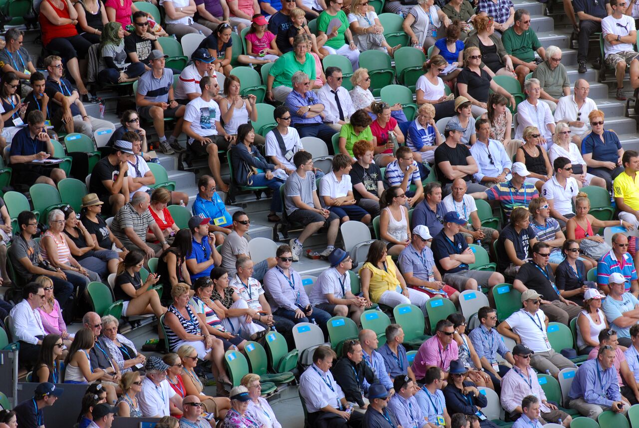 Spectators fill rows of green and white seats at the Australian Open, many in sunglasses and hats, watching the match.