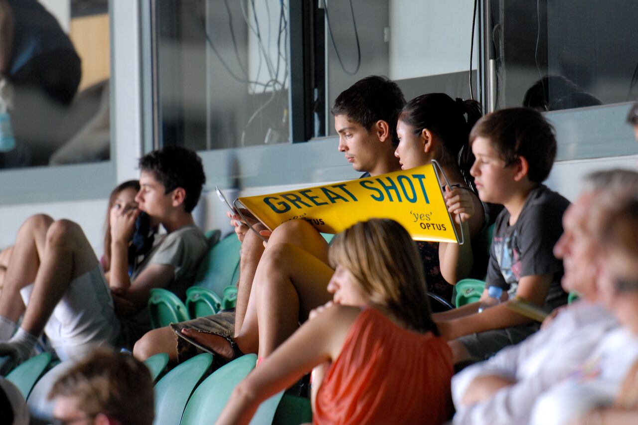 Young spectators sit in green seats at the Australian Open, one holding up a yellow "Great shot" sign from Optus.