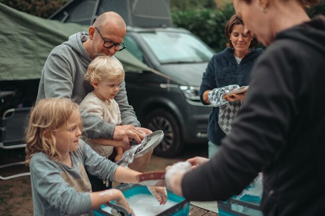 A family washing dishes together in soapy basins at a campsite, with a van in the background.
