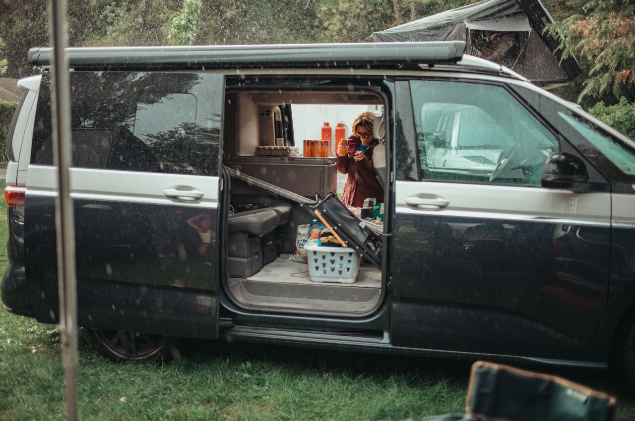 A woman prepares drinks inside a camper van with its sliding door open on a rainy day.