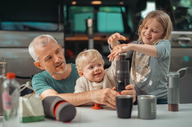 A man and two young children make coffee with a portable press at a table in front of a camper van.