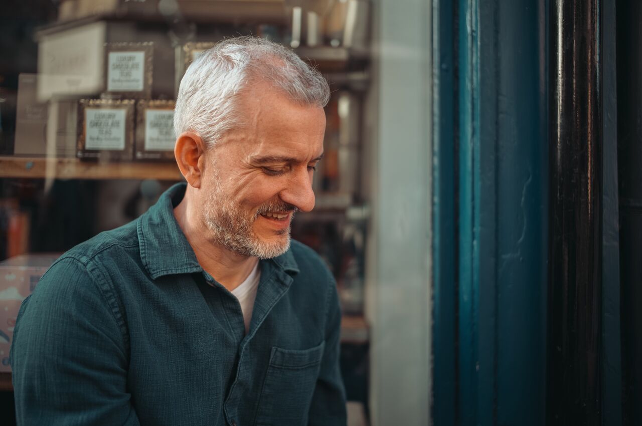 A smiling man with gray hair looking down, outside a shop window.