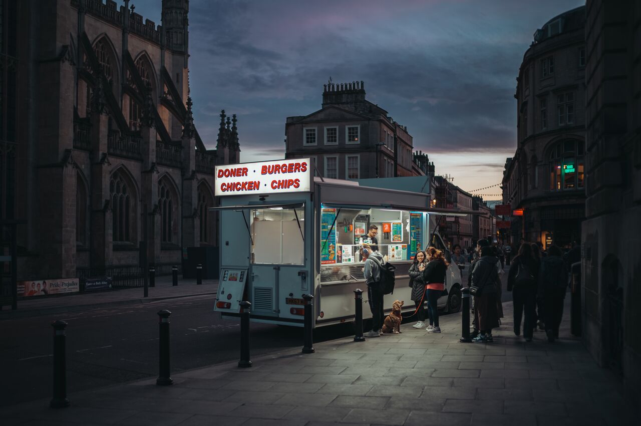 A food truck parked beside Bath Abbey, serving doner, burgers, and chips to a small crowd at dusk.