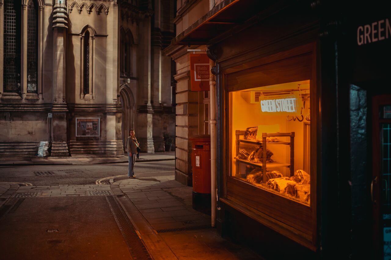 A lit butcher shop window at dusk, with a woman waiting on the street beside a historic church.