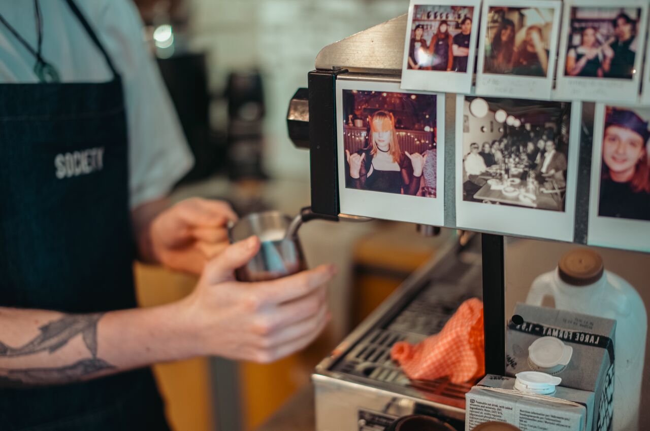 A barista pouring milk at an espresso machine decorated with Polaroid photos.