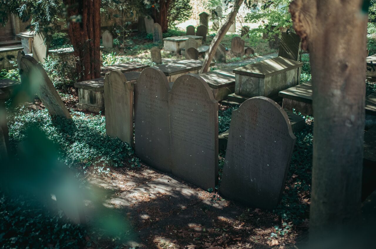 Old gravestones in a shaded cemetery, surrounded by trees and greenery.
