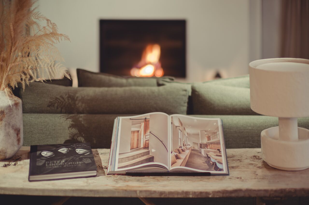 An open design book on a rustic wooden table, in front of a green sofa, with a warm fireplace in the background.