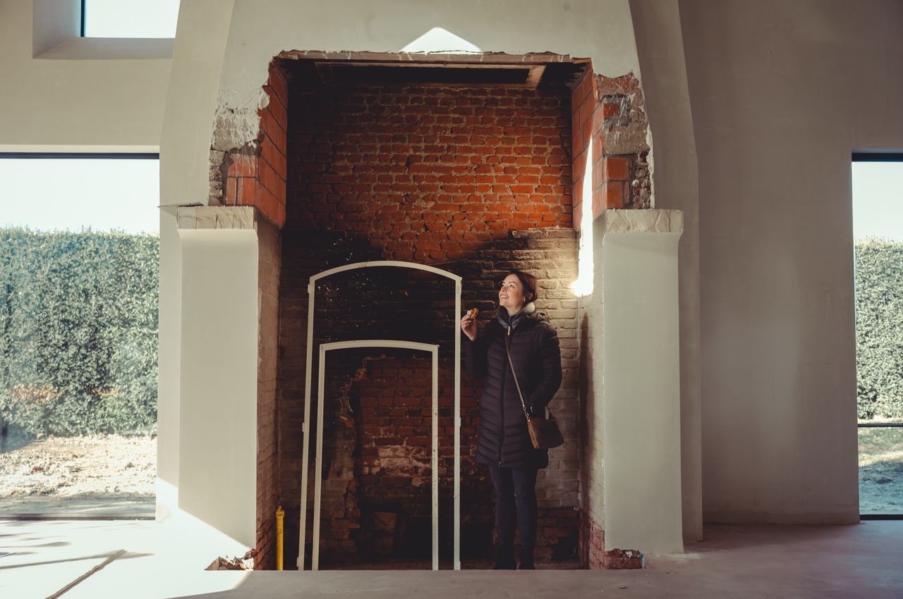 A woman is standing inside a large fireplace that is currently being rebuild. She is enjoying a baked treat while taking in the scene around her.
