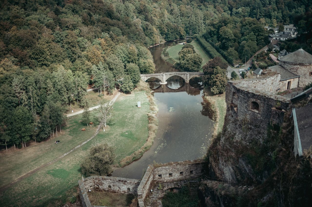 Castle walls rising above a river with a medieval bridge and green banks..