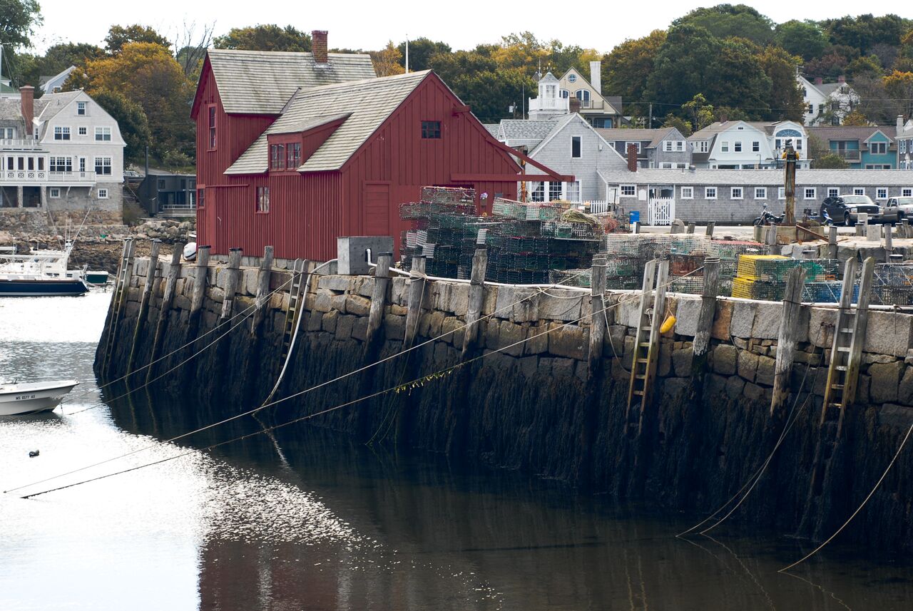 A red wooden building stands by a stone pier with stacked lobster traps and moored boats in the harbor.