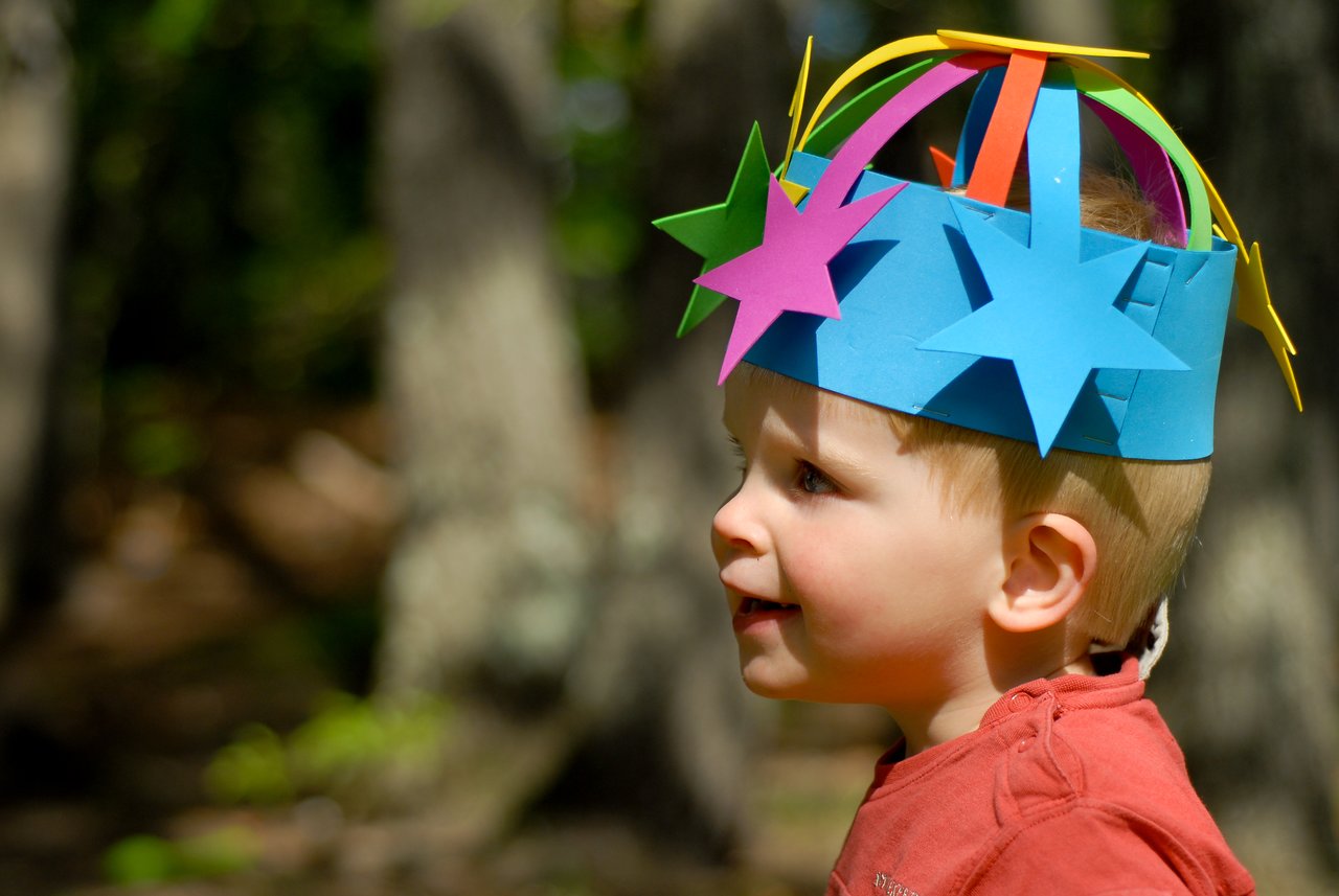 A young child wearing a colorful paper crown with stars smiles while looking to the side at a party.