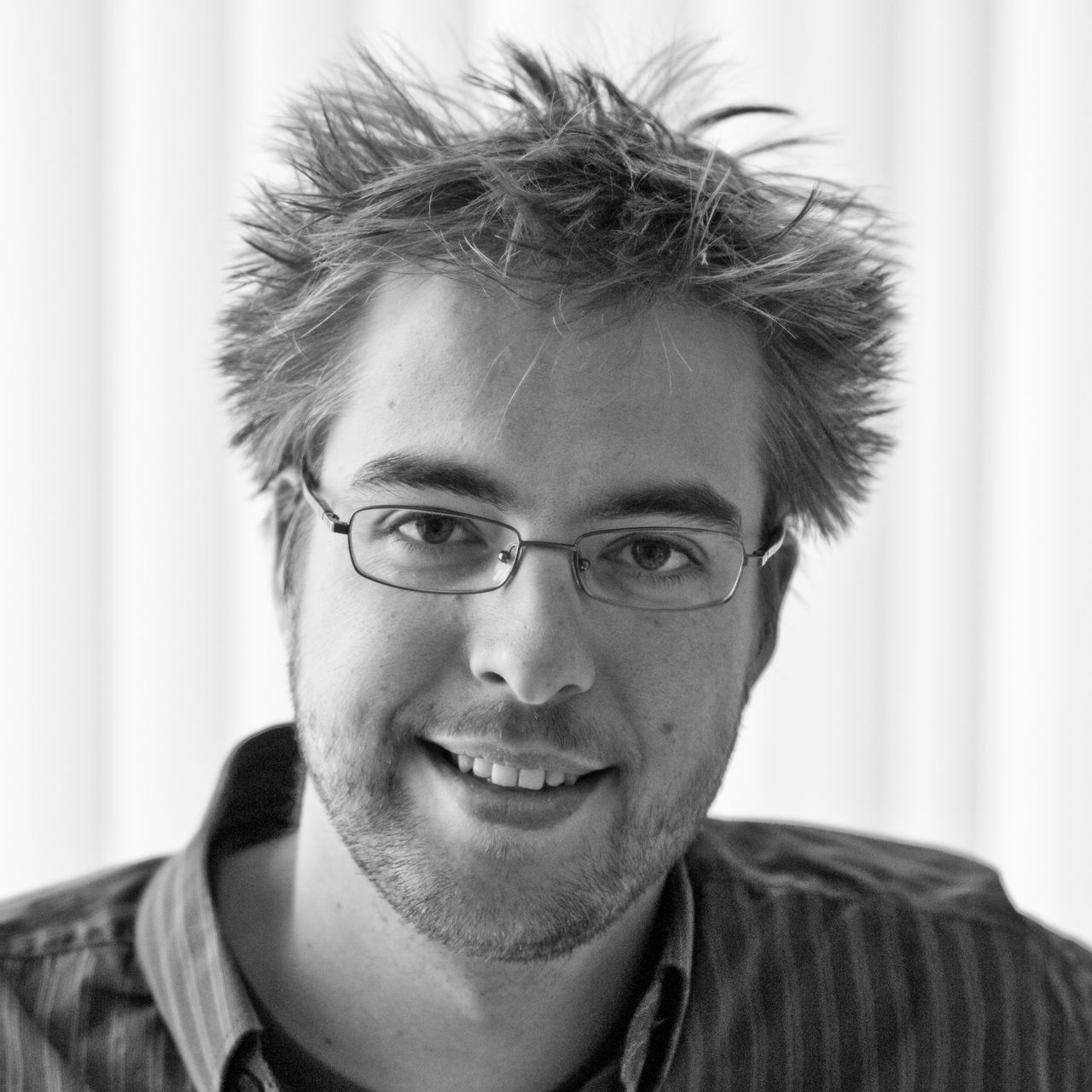 Black and white portrait of a man with glasses and messy hair, smiling at the camera.