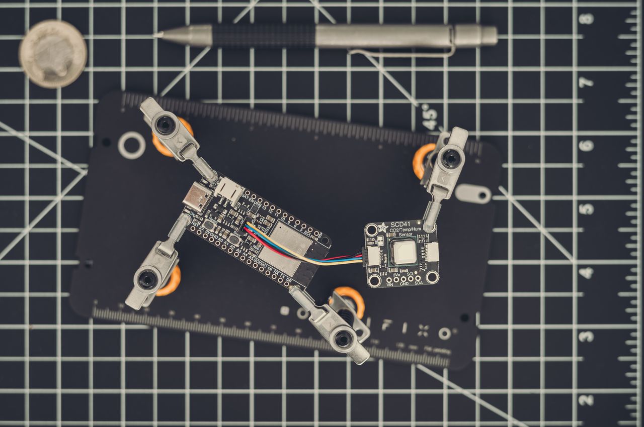 An ESP32-S3 development board is linked to an SCD41 CO2 and temperature sensor. For scale, a coin and pen are included. The SCD41 sensor is roughly equivalent in size to the coin, and the ESP32-S3 board is about twice the coin's diameter.