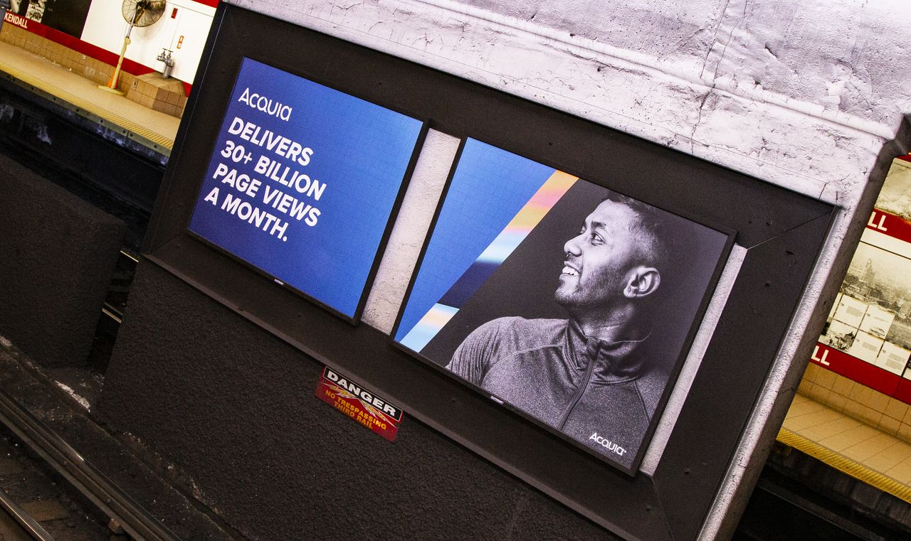 Two large Acquia advertisements at Kendall Square station, one showing text about page views and the other a smiling man.