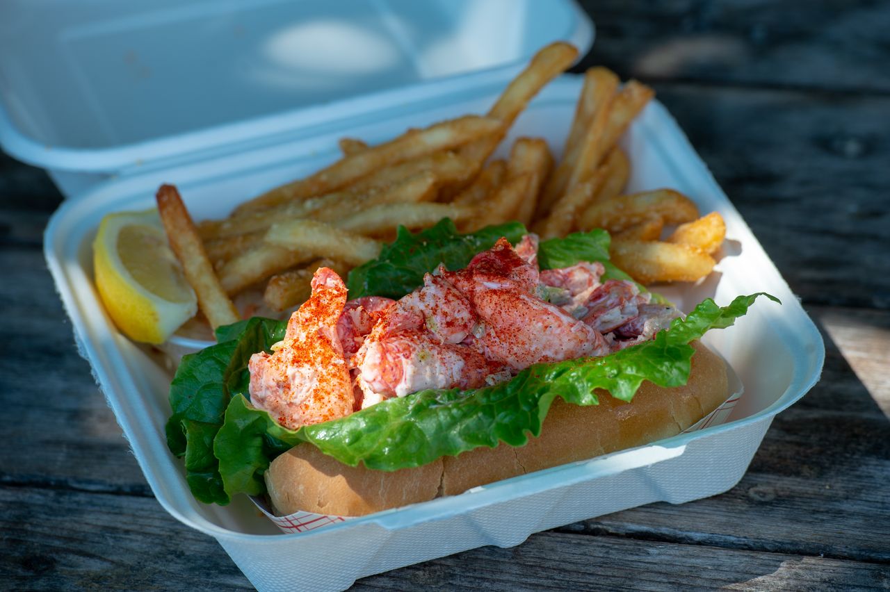 A lobster roll with lettuce and seasoning, served with fries and a lemon wedge in a foam container.