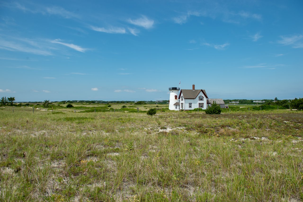 A white lighthouse with an attached house stands in an open grassy field under a clear blue sky.