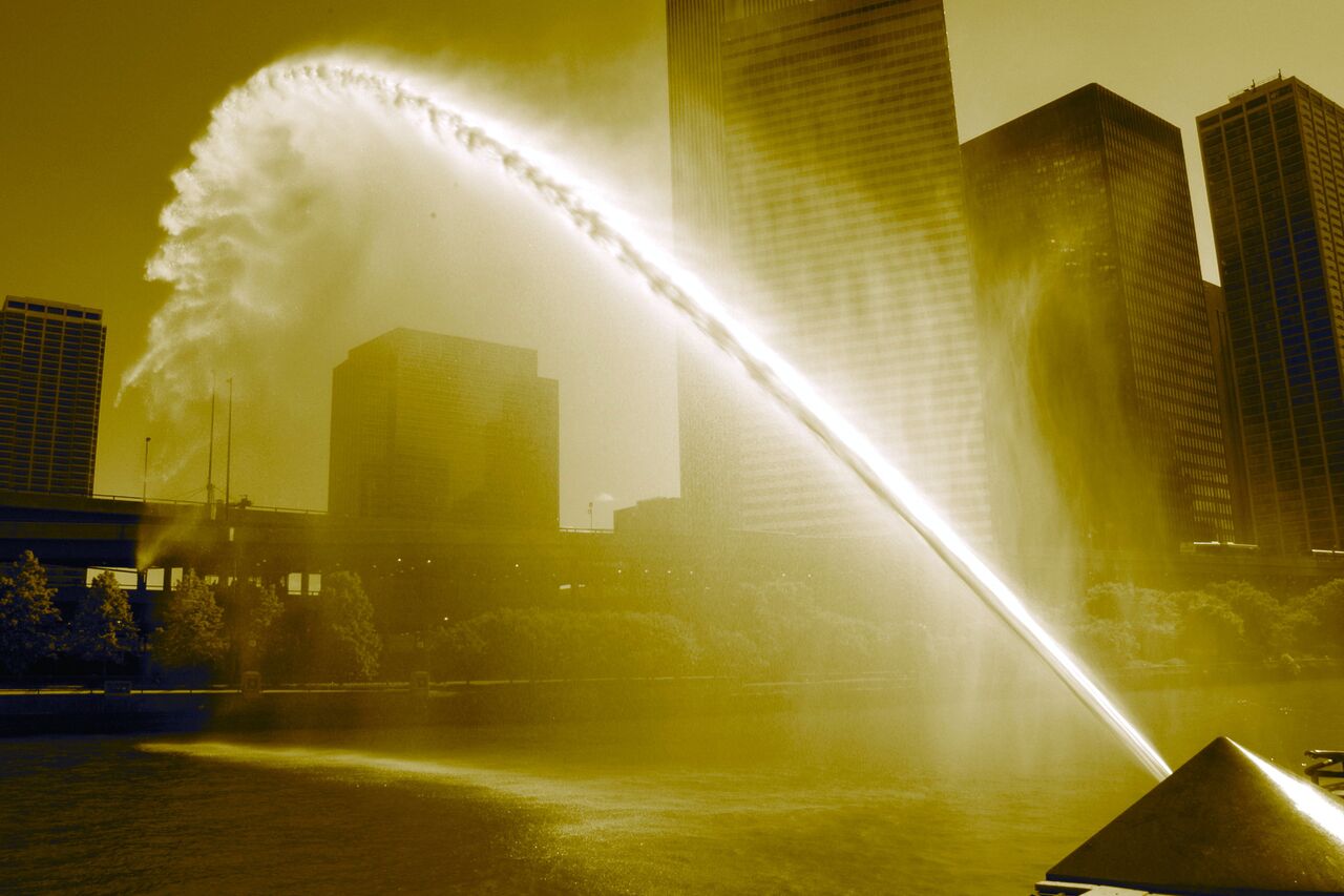 A large fountain sprays an arc of water over a river, with tall buildings in the background.