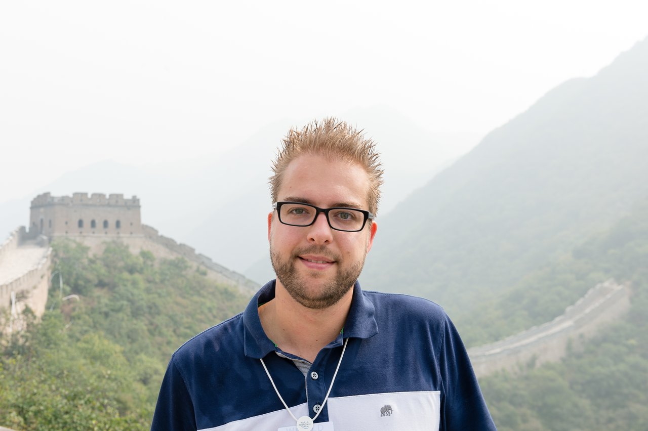 A man wearing glasses and a blue shirt stands in front of the Great Wall of China.