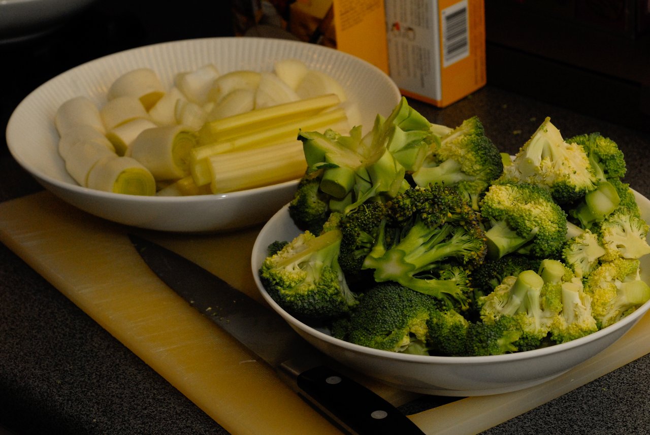 Chopped broccoli and leeks in bowls on a cutting board, with a knife nearby, prepared for cooking.