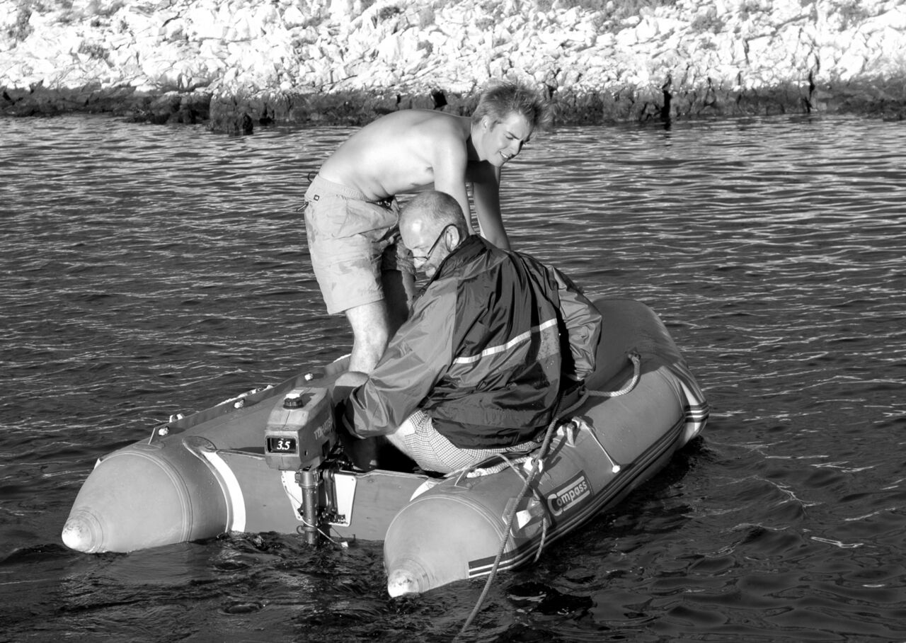 One person adjusts the motor while another watches on a small inflatable boat in the water.