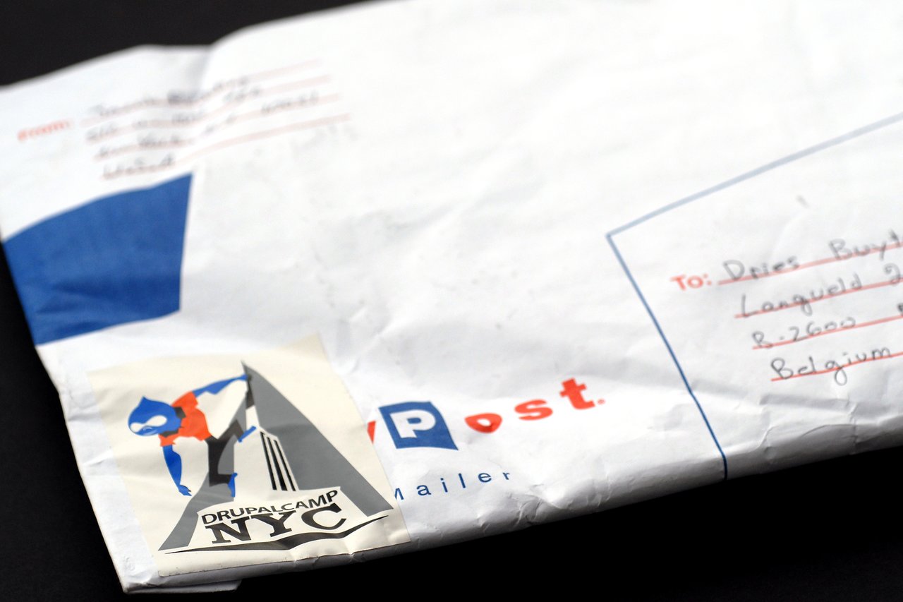 A mailing envelope with a Drupalcamp NYC sticker and handwritten address details, placed on a dark surface.