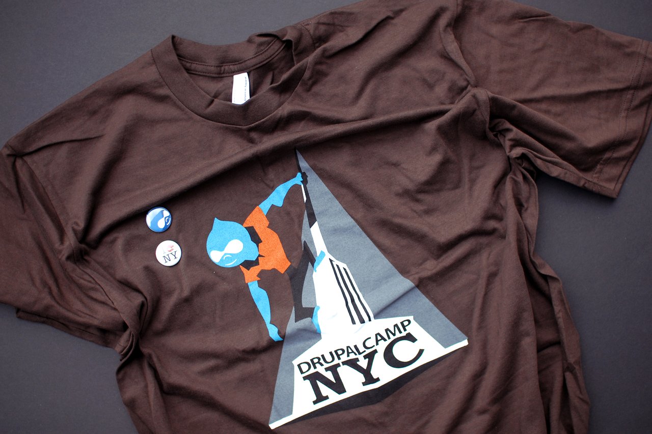 Brown Drupalcamp NYC T-shirt with a graphic of a climber and two Drupal-themed buttons on fabric.