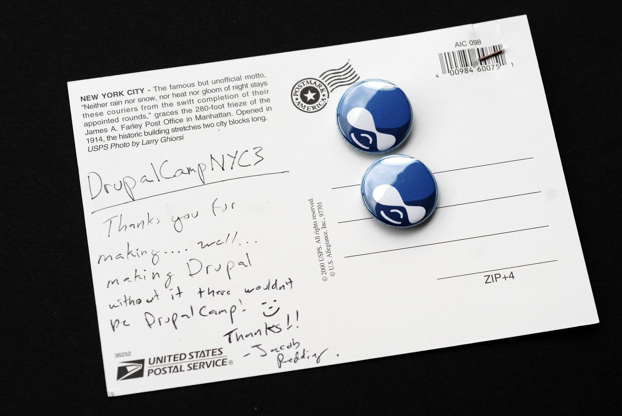 A postcard with a handwritten message about DrupalCamp NYC and two blue Drupal logo buttons placed on it.