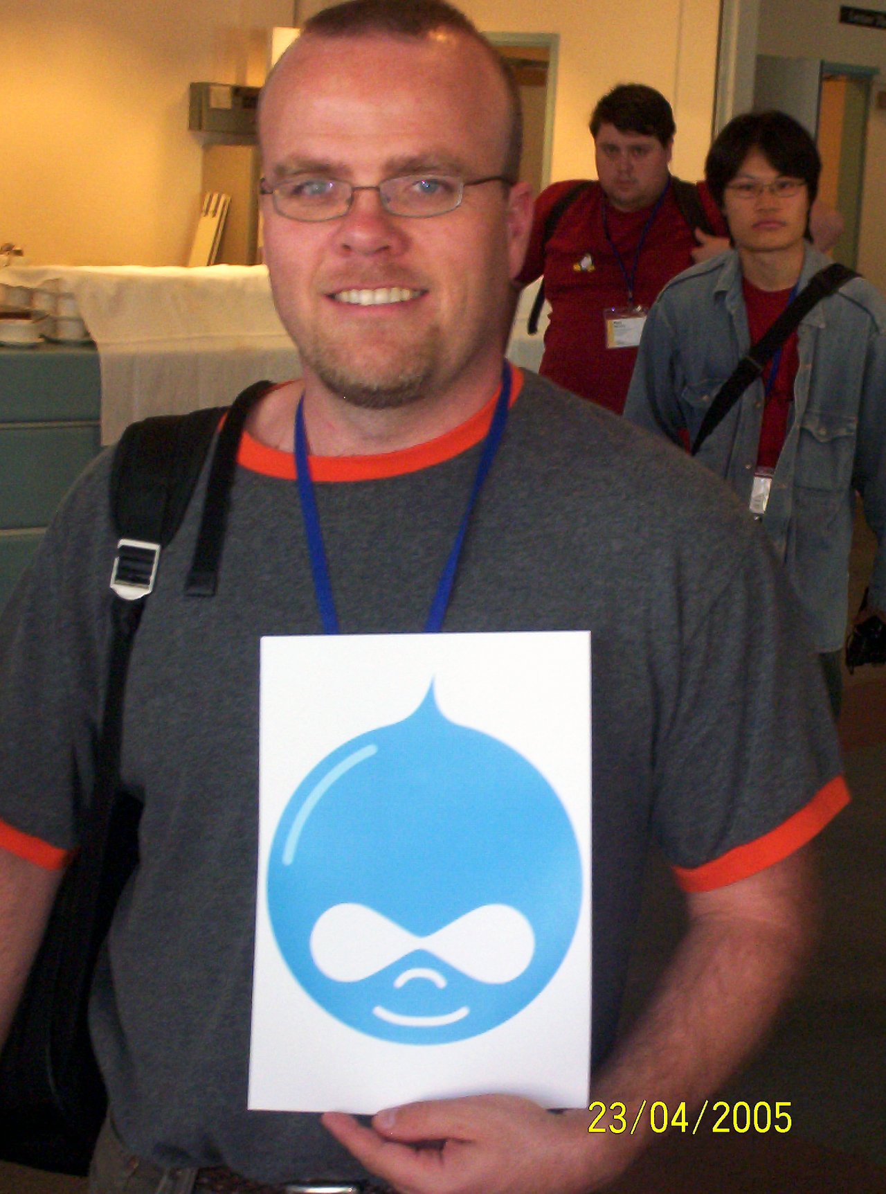 A man wearing glasses holds a printed Drupal Druplicon logo while smiling at the camera.