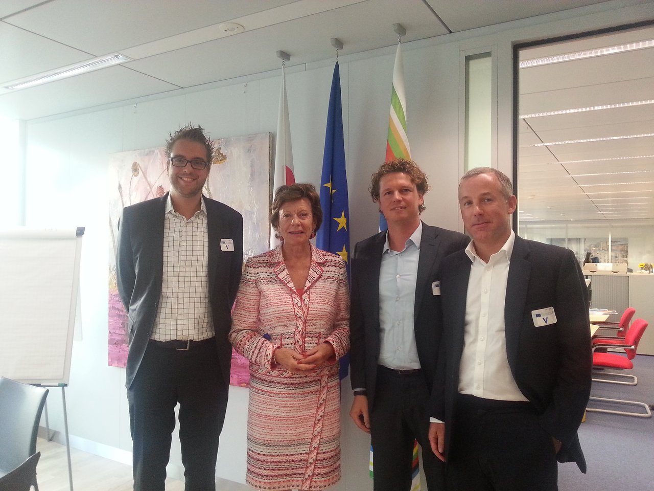Four people stand together in an office, posing for a group photo in front of flags.